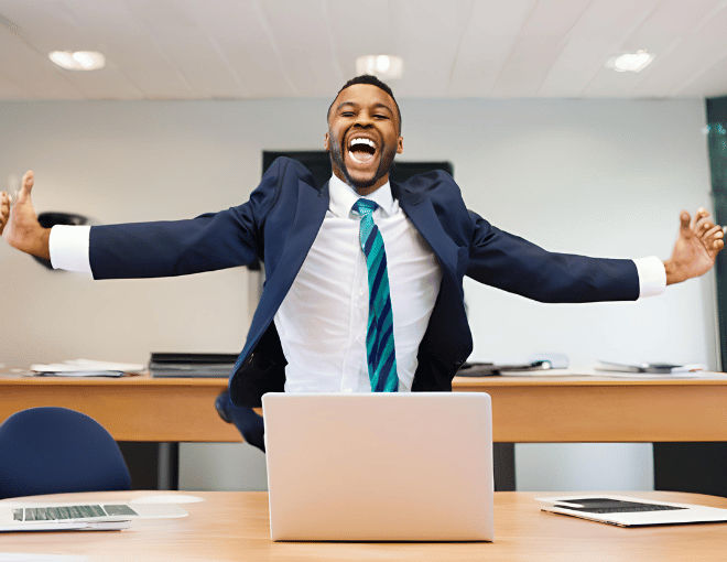 A businessman jumps with joy, arms raised, wide smile, laptop open on desk, suit and tie immaculate, suggesting a huge success or windfall.