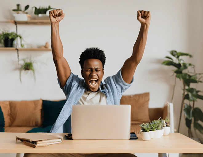 A man sits jubilantly at his laptop, fists raised in victory.
